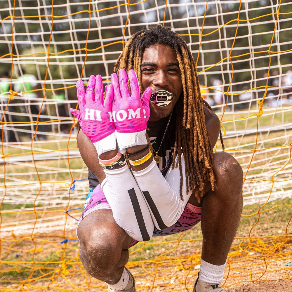 Athlete wearing Cutters Rev Pro 5.0 Hi Mom LE Football Receiver Gloves in pink, kneeling on field with gloves raised to show 'HI MOM!' text design.