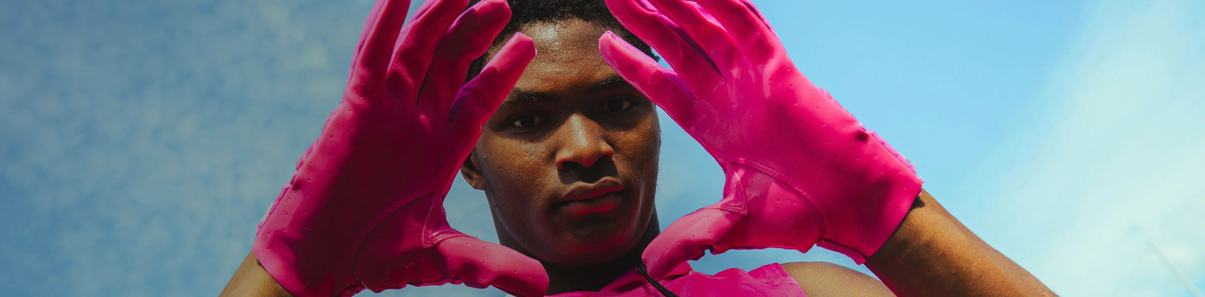 Header image for the 'Pink Football Gloves' Collection by Cutters Sports. A football player frames their face with both hands, showing off bright pink receiver gloves against a clear blue sky.