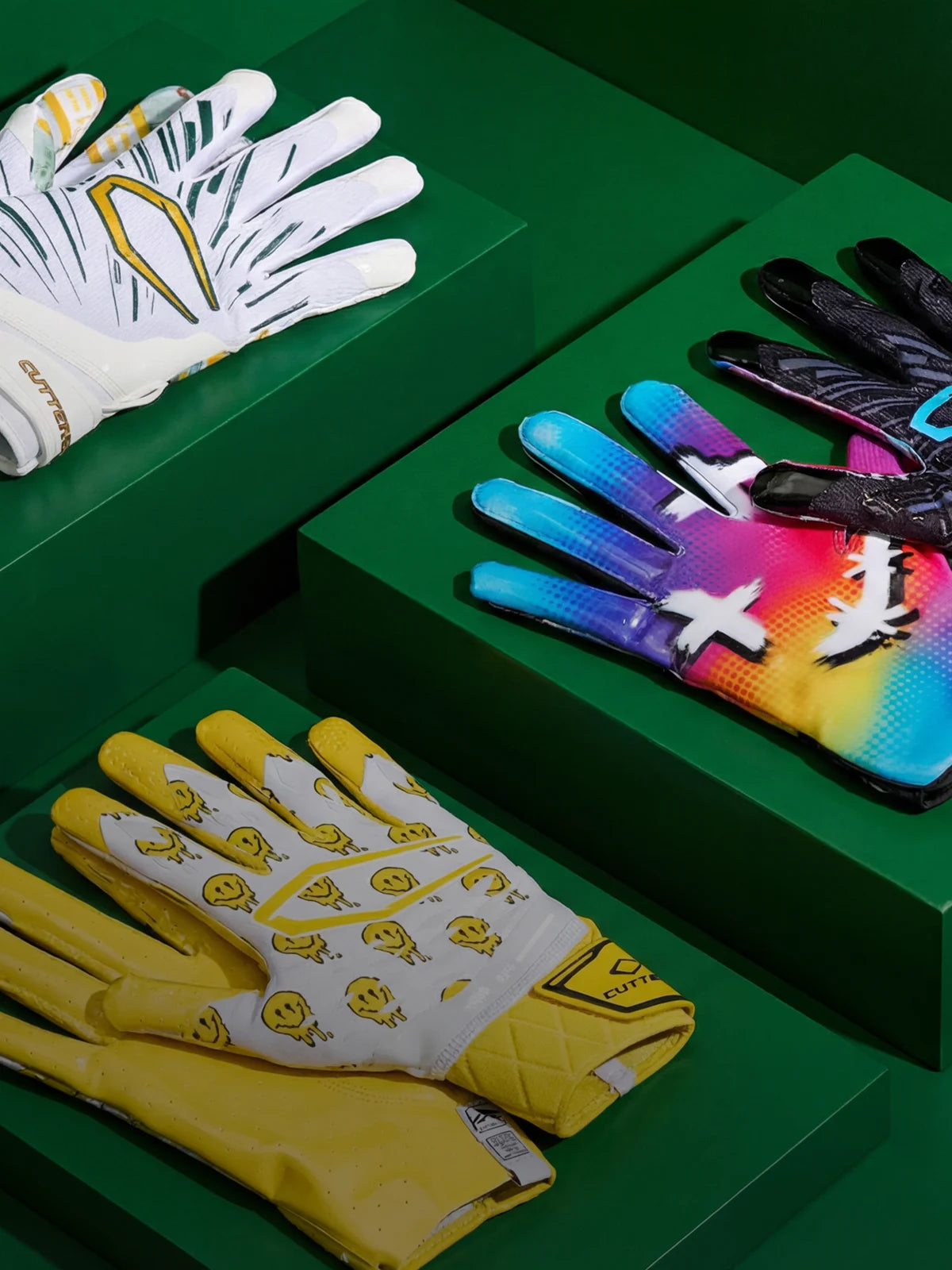 Four pairs of gloves on a green surface, including white with yellow accents, colorful, yellow with smiley face pattern, and black with blue accents.