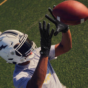Football receiver reaching overhead to catch a pass wearing black Cutters Rev Pro 7.0 Solid receiver gloves on a turf field