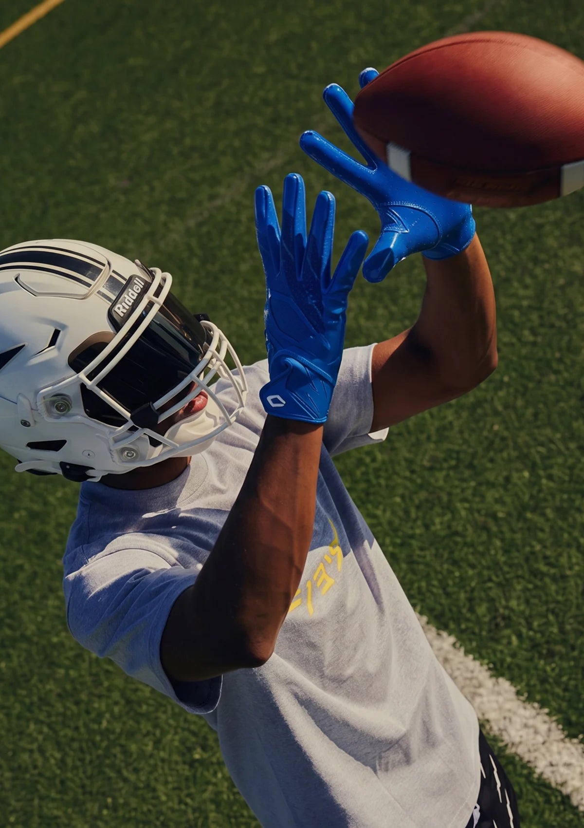 A football player in a white helmet and light gray practice shirt reaches for a brown football on a green turf field, wearing bright blue gloves.
