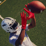 Football player reaching up to catch a pass wearing red Cutters Rev Pro 7.0 Solid receiver gloves on a turf field