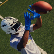 Receiver in a white helmet catching a football wearing royal blue Cutters Rev Pro 7.0 Solid receiver gloves on a turf field