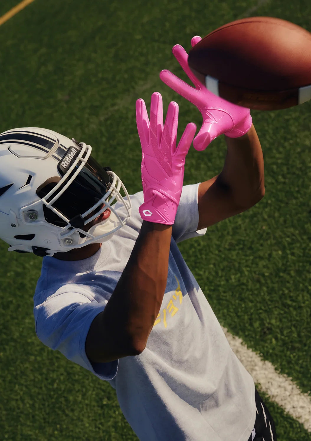 Close-up overhead view of a football player in a white helmet reaching up to catch a football while wearing pink Cutters Rev Pro 7.0 receiver gloves.