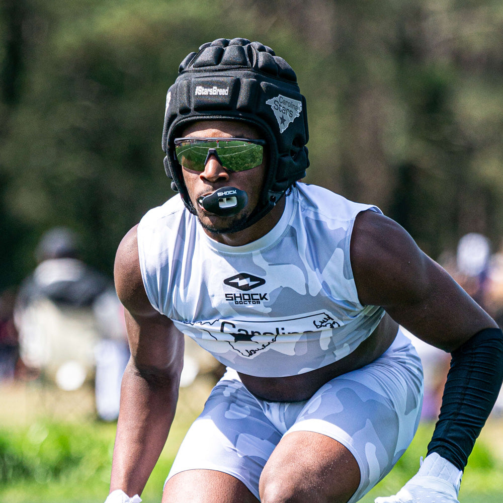 Football player in white camo uniform wearing a black Shock Doctor Max AirFlow Lip Guard, padded headgear, and sunglasses in game stance.